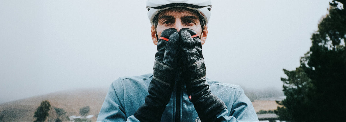 A man wearing a white helment, a blue cycling jacket, and black winter gloves  with a rainy outdoor landscape in the background.