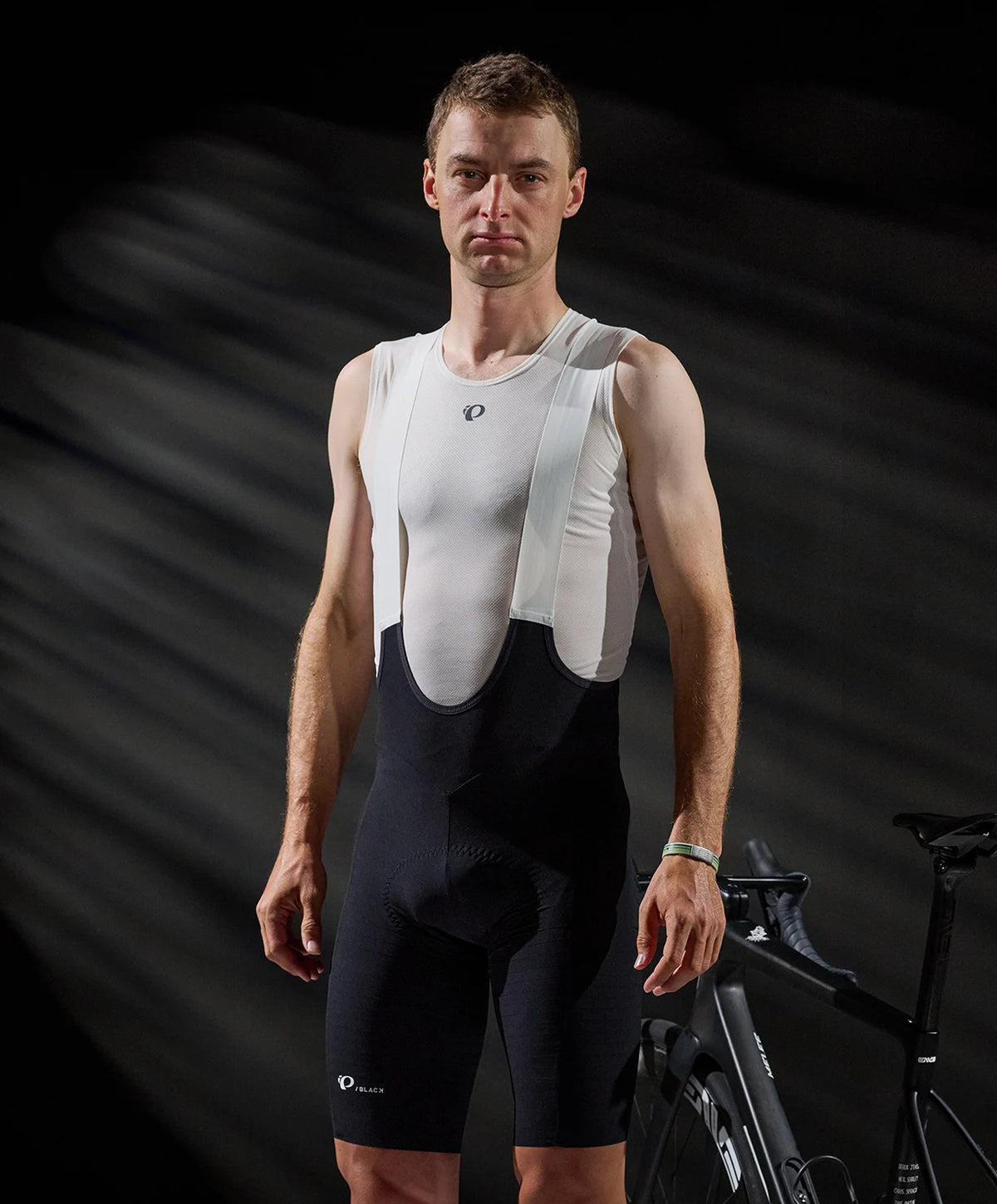 Man wearing a white cycling jersey and black bib shorts on a dark background