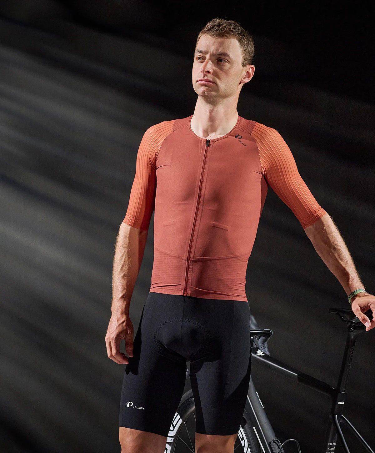 Man wearing a red cycling jersey and black shorts standing next to a bicycle on a dark background