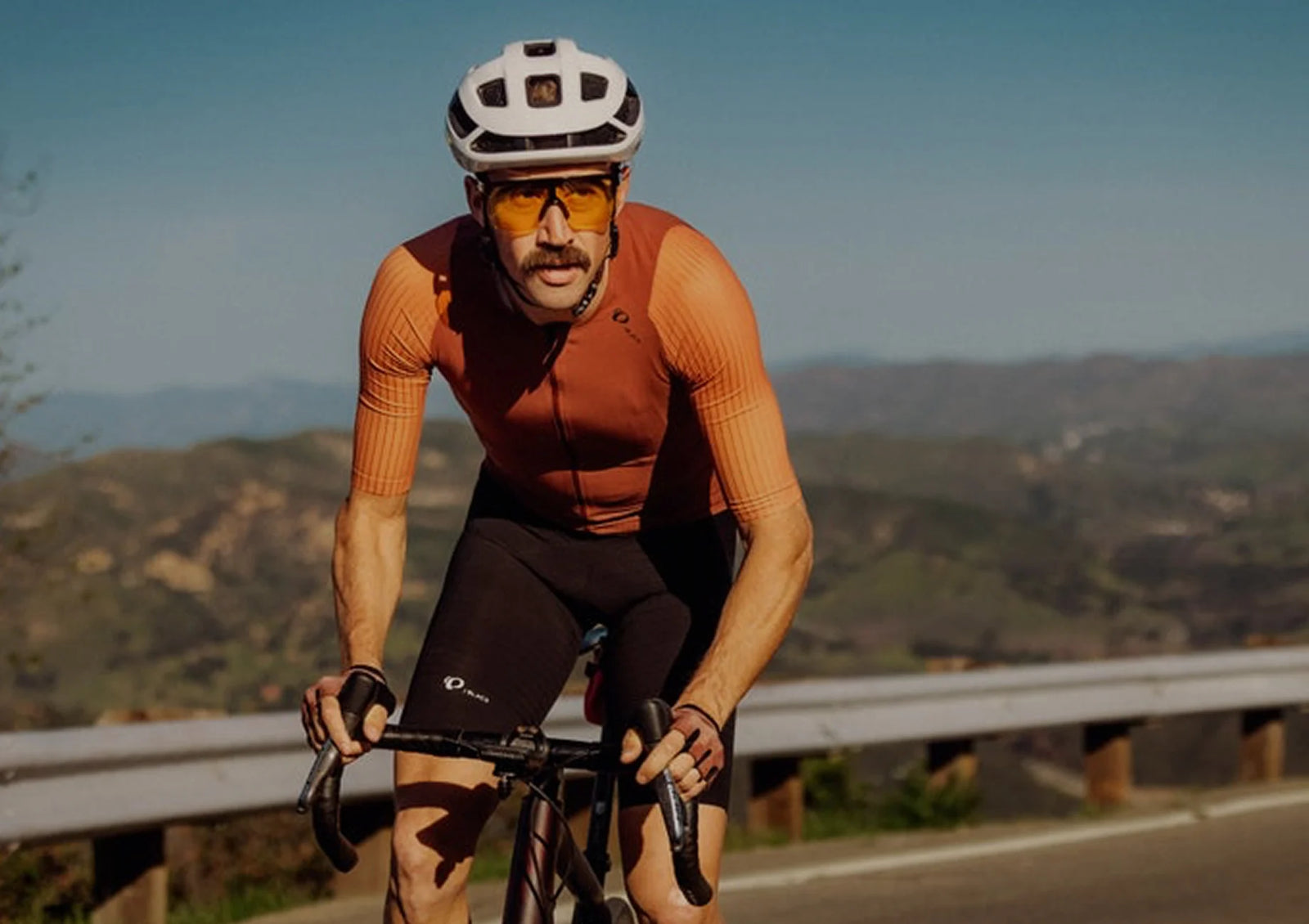 Cyclist in orange jersey and black shorts riding on a road with mountains in the background