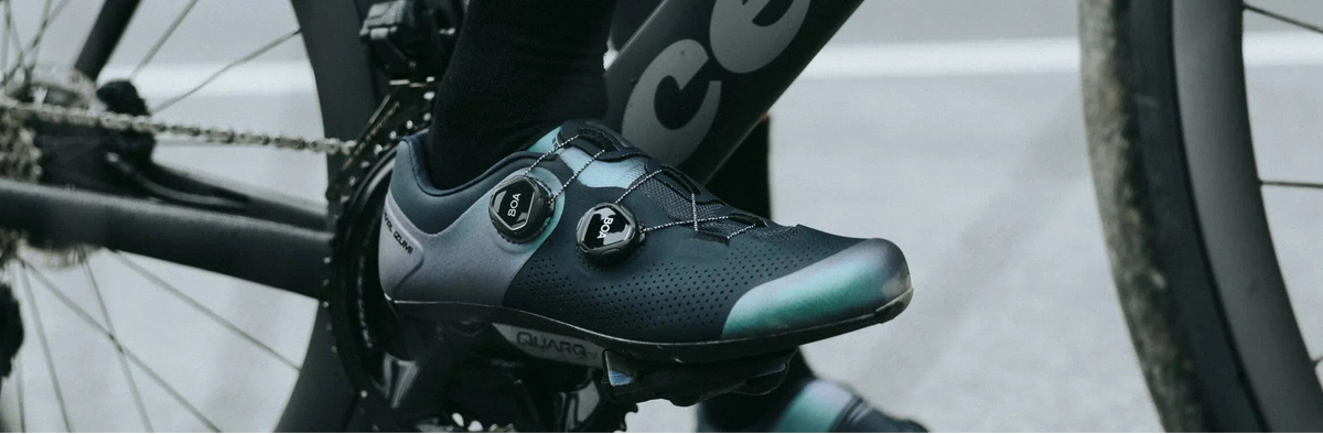 A close-up of a cyclist wearing a black road cycling shoe with teal accents, mounted on a bike pedal.