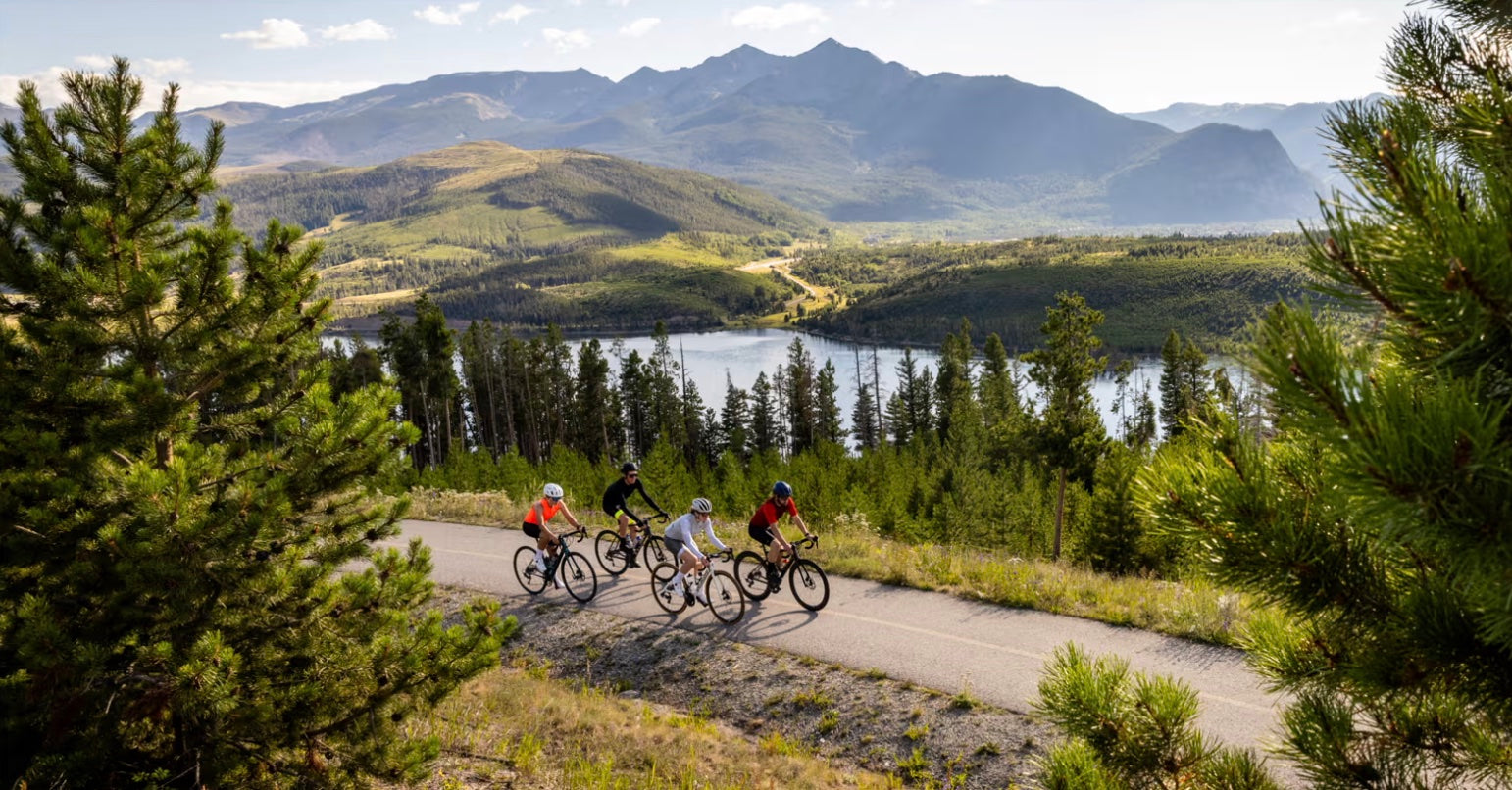 Four cyclists on a scenic mountain trail with a lake and mountains in the background